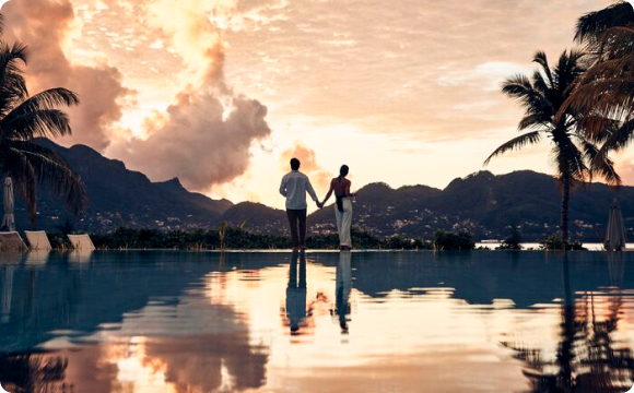 Couple debout au bord d’une piscine face à un coucher de soleil, avec des palmiers et des montagnes en arrière-plan.